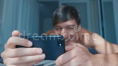 快乐微笑的男人在家里躺在床上看智能<strong>手机</strong>上的<strong>视频</strong>。 裸笑男用智能<strong>手机玩</strong>游戏